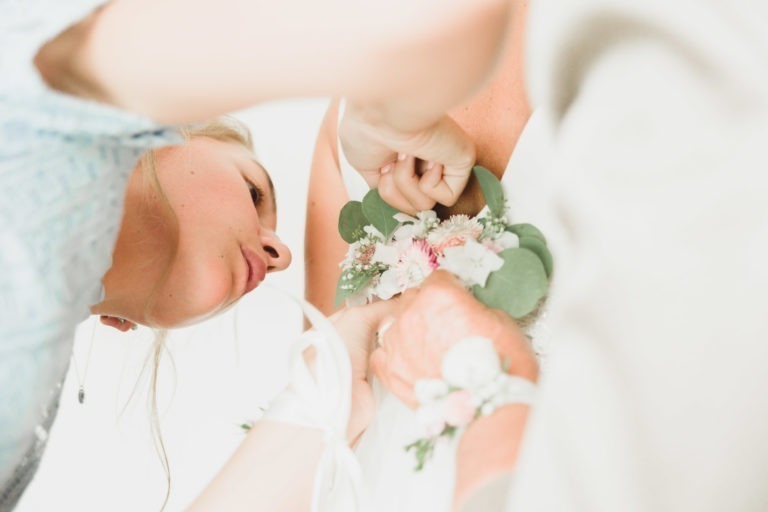 Une image en gros plan montre une femme en train d'ajuster un bouquet de fleurs au poignet d'une autre personne. L'accent est mis sur les mains et le bouquet, qui présente des fleurs blanches et roses avec des feuilles vertes. L'arrière-plan est légèrement flou, soulignant l'interaction détaillée et la simplicité de cet acte lors d'un mariage.