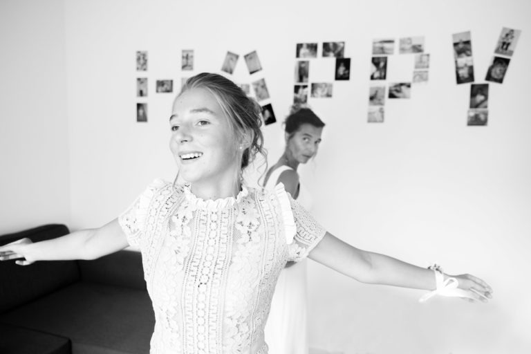 Une photo en noir et blanc de deux femmes, probablement en train de célébrer un moment. La femme au premier plan, avec les cheveux tressés et une robe en dentelle, a les bras tendus et sourit. La femme à l'arrière-plan sourit également. Derrière elles, le mot « HAPPY » est écrit à l'aide de diverses photos accrochées au mur, capturant une scène de simplicité et de générosité.