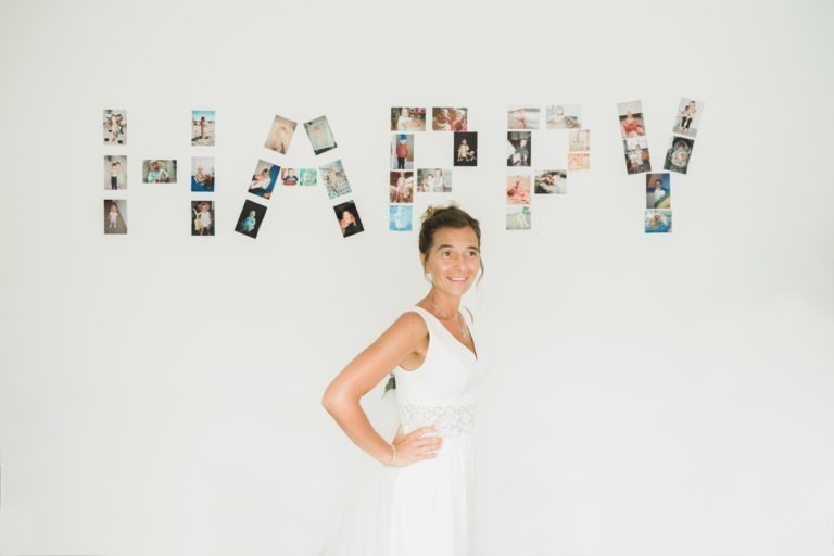 Une femme en robe blanche sans manches se tient debout, souriante, la main sur la hanche, rayonnant de simplicité. Le mur du fond porte le mot « HAPPY » écrit en grandes lettres, chacune composée de petites photos individuelles. La scène est bien éclairée et dégage une atmosphère minimaliste et lumineuse.