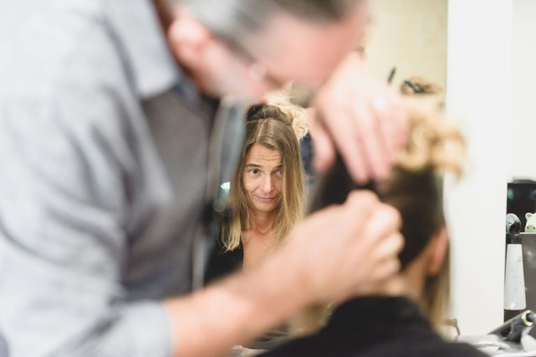 Une femme aux cheveux blonds mi-longs se regarde dans le miroir avec un léger sourire tandis qu'un coiffeur s'occupe de ses cheveux. Le coiffeur, qui porte des lunettes et une chemise grise, se concentre sur ses cheveux, qui sont relevés ou coiffés avec une élégante simplicité. L'arrière-plan est un salon de coiffure lumineux et propre.