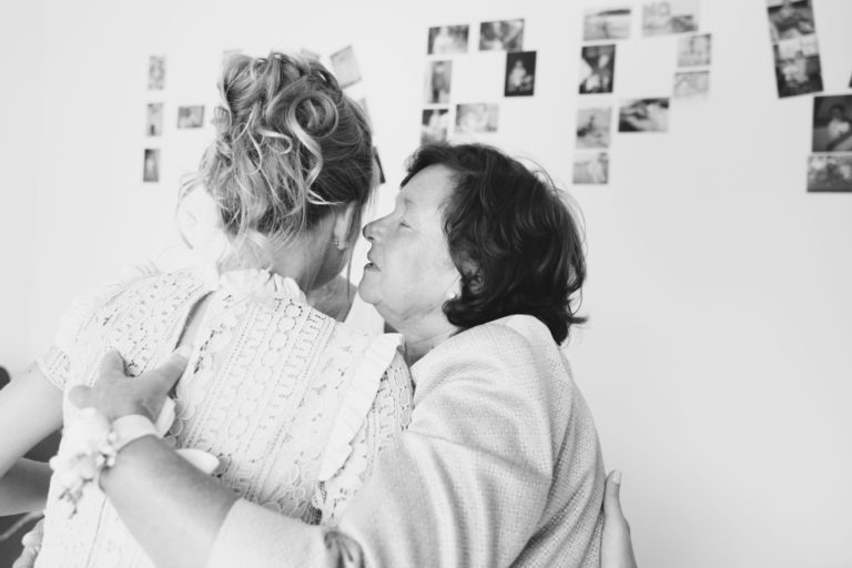 Une photo en noir et blanc capturant un moment intime entre deux femmes qui s'embrassent. La femme de gauche, avec les cheveux bouclés relevés et vêtue d'une robe en dentelle, tourne le dos à l'objectif. La femme de droite, aux cheveux courts, ferme les yeux dans un simple geste d'affection pendant qu'elles s'embrassent. Les photos en arrière-plan ornent le mur.