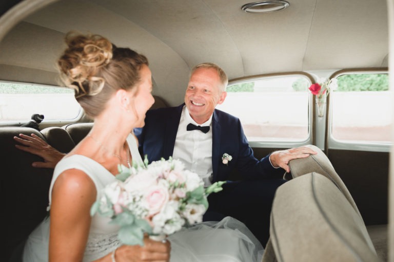 Un couple de mariés est assis à l'arrière d'une voiture ancienne, illustrant la simplicité de l'amour. La mariée, vêtue d'une robe de mariée blanche avec un chignon, tient un bouquet de fleurs. Le marié, vêtu d'un costume sombre avec un nœud papillon et une boutonnière, lui sourit joyeusement alors qu'ils célèbrent leur mariage.