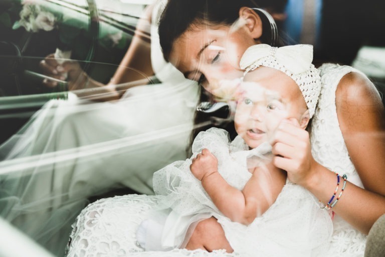 Une jeune fille berce avec amour un nourrisson vêtu de blanc, qui porte un bandeau avec un gros nœud. Ils ont tous deux les cheveux noirs et sont vêtus de vêtements blancs délicats. La scène, empreinte d'un air de simplicité, semble capturée à travers une fenêtre réfléchissante, ajoutant une qualité douce et surréaliste à l'image.