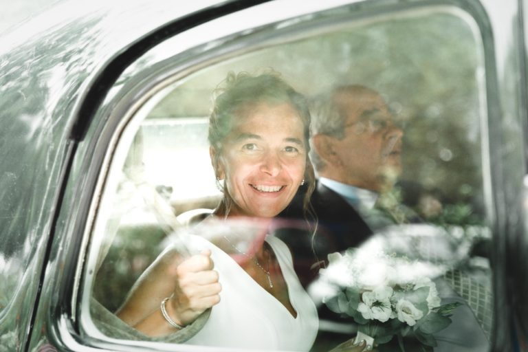 Une femme souriante, vêtue d'une robe blanche, est assise dans une voiture, tenant le cadre de la fenêtre. À côté d'elle, un homme âgé portant des lunettes et un costume est assis, regardant devant lui. La femme porte un bouquet de fleurs blanches sur ses genoux, symbolisant la simplicité de leur moment de complicité. Tous deux sont visibles à travers la vitre légèrement réfléchissante de la voiture.