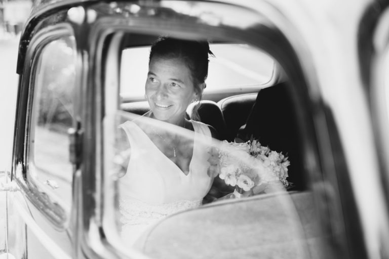 Une photo en noir et blanc d'une femme souriante en robe de mariée assise sur le siège arrière d'une voiture ancienne. Elle tient un bouquet de fleurs et regarde par la fenêtre ouverte avec un air de simplicité sereine. L'image capture un moment candide et joyeux le jour de son mariage.