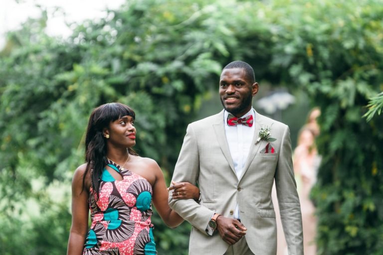 Un couple marche bras dessus, bras dessous dans un jardin luxuriant. La femme porte une robe à motifs colorés et l'homme est en costume gris clair avec un nœud papillon rose et une boutonnière. Un feuillage vert se courbe au-dessus de leur tête, créant une voûte naturelle, alors qu'ils partagent ce mariage émouvant et touchant. Une silhouette floue apparaît à l'arrière-plan.