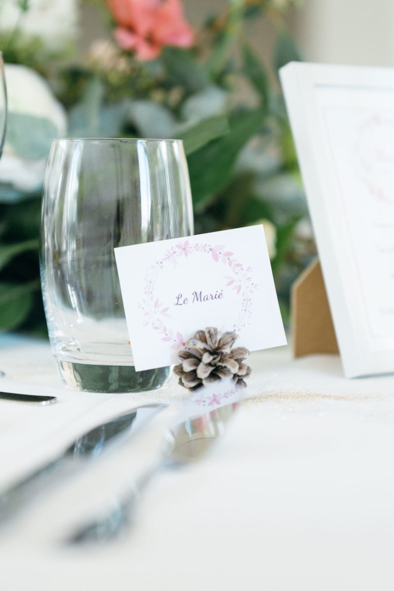 Une table de mariage avec verres, couverts et marque-place pliée décorée d'une bordure florale violette et du texte « Le Marié ». Le marque-place est maintenu en place par une petite pomme de pin. L'arrière-plan flou comprend de la verdure et des fleurs roses, capturant l'essence d'un mariage émouvant et touchant.