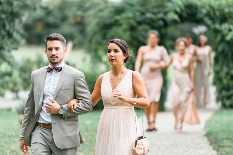 Un homme en costume gris clair avec un nœud papillon accompagne une femme en robe rose pâle le long d'une allée de jardin lors d'un mariage émouvant et touchant. Ils regardent devant eux en marchant. Derrière eux, un groupe de femmes portant des robes de couleur claire similaires les suit. L'arrière-plan est luxuriant et vert avec une arche feuillue.