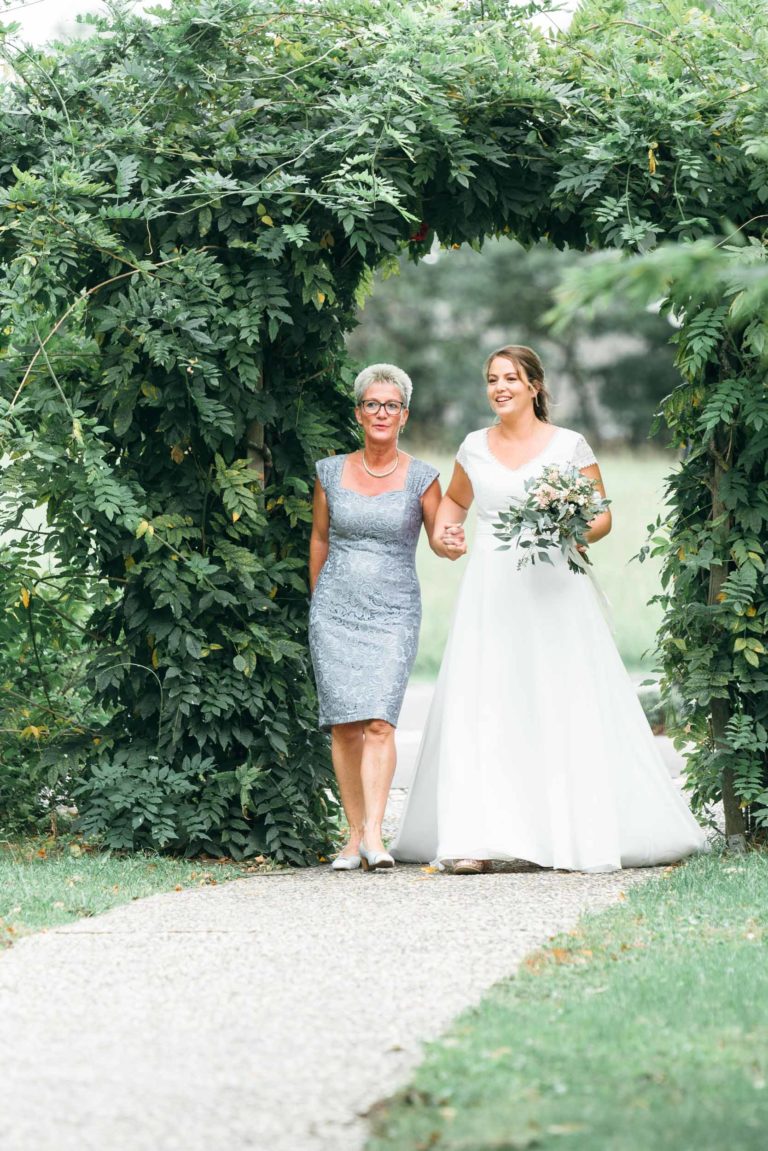 Une mariée en robe blanche et voile marche sur une allée, tenant un bouquet de fleurs et bras dessus bras dessous avec une femme plus âgée en robe argentée. Elles passent sous une arche verte luxuriante couverte de feuilles et de vignes, souriant joyeusement à ce mariage émouvant et touchant. Le décor est en extérieur, avec de la verdure en arrière-plan.