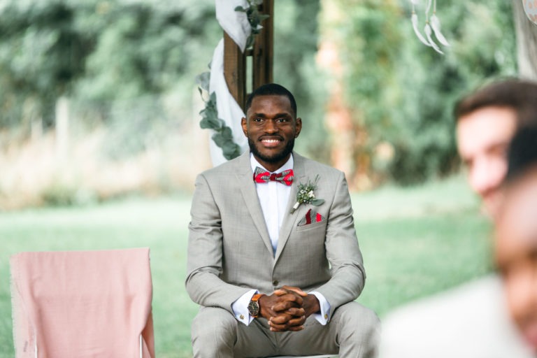 Un homme est assis dehors, souriant et vêtu d'un costume gris clair avec une chemise blanche et un nœud papillon rouge. Il tient ses mains jointes, avec une boutonnière épinglée sur son revers gauche, complétée par une pochette rouge. Le fond de verdure floue et de décorations partiellement visibles ajoute à l'atmosphère de ce mariage émouvant et touchant.