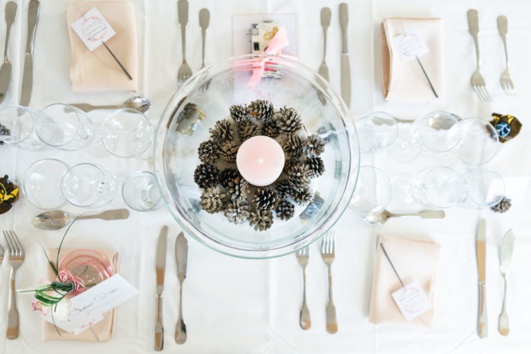 Une table bien dressée vue d'en haut pour un mariage émouvant et touchant. La pièce maîtresse est un bol rempli de pommes de pin et une seule bougie blanche. La table est recouverte de linge blanc, de serviettes couleur pêche, de couverts en argent, de verres à vin et de petites cartes sur les serviettes. Des rubans roses et des décorations en fil de fer vert sont également présents.