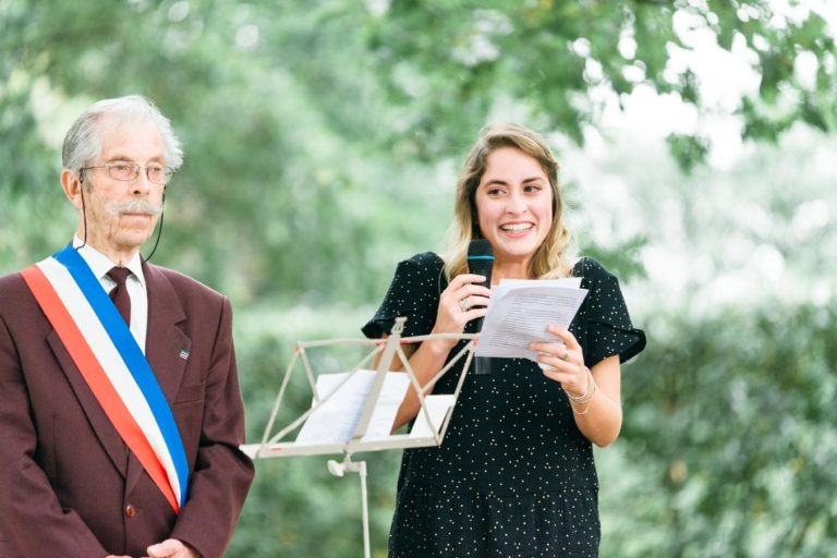 Une jeune femme en robe noire parle dans un micro tout en tenant une feuille de papier. À côté d'elle, un homme plus âgé aux cheveux gris et à la moustache, portant des lunettes et un costume marron avec une ceinture tricolore, se tient sereinement. Ils sont dehors au milieu d'un feuillage vert luxuriant, comme s'ils capturaient l'essence d'un mariage émouvant et touchant.