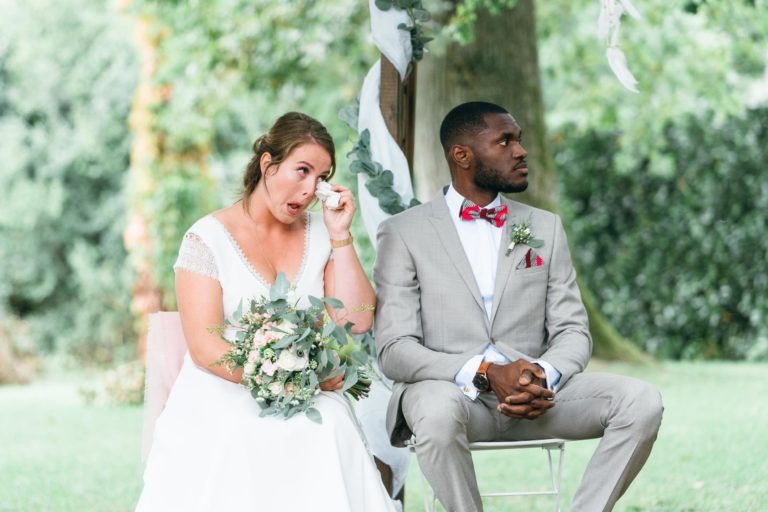 Une mariée en robe blanche, tenant un bouquet et essuyant une larme, est assise à côté du marié en costume gris clair avec un nœud papillon rouge et une pochette. Tous deux sont assis sous un auvent extérieur décoré de verdure, avec un arbre et un arrière-plan luxuriant. Le marié regarde au loin pendant ce mariage émouvant et touchant.