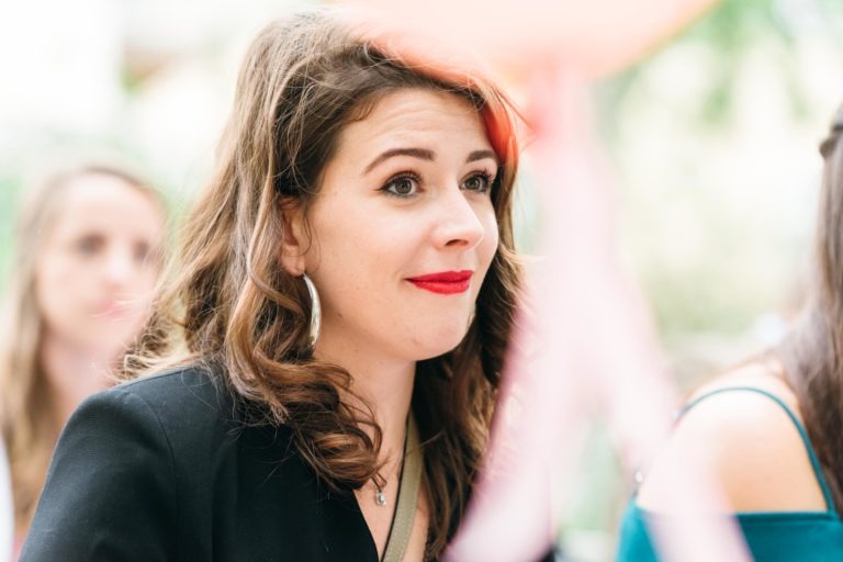 Une femme aux cheveux bruns ondulés et au rouge à lèvres rouge vif est vue en gros plan. Elle porte un haut noir et de grandes boucles d'oreilles créoles, et semble être à l'extérieur, peut-être à un mariage émouvant et touchant, avec un arrière-plan flou laissant entrevoir d'autres personnes et de la verdure. L'expression de son visage est celle d'une agréable surprise.