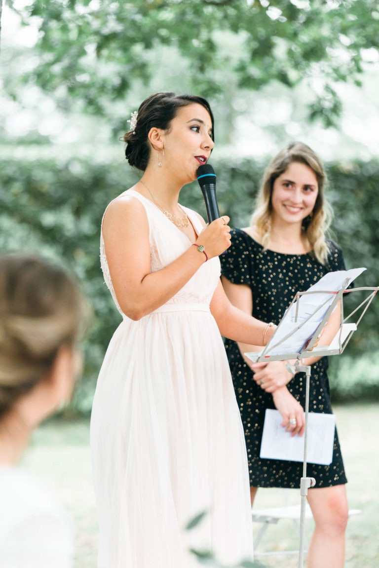 Une femme en robe rose pâle parle dans un micro tout en tenant des papiers, dans un décor extérieur verdoyant. À proximité, une autre femme en robe noire à pois blancs sourit et tient un carnet. La scène dégage un air de mariage émouvant et touchant.