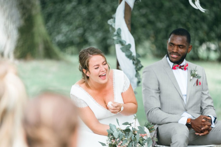 Une mariée en robe blanche aux manches en dentelle rit de bon cœur tout en tenant un petit objet dans sa main. Elle est assise à côté d'un marié en costume gris clair avec un nœud papillon rouge, tous deux assis à l'extérieur. L'arrière-plan montre de la verdure et un arrangement floral drapé, capturant l'essence de ce mariage émouvant et touchant.