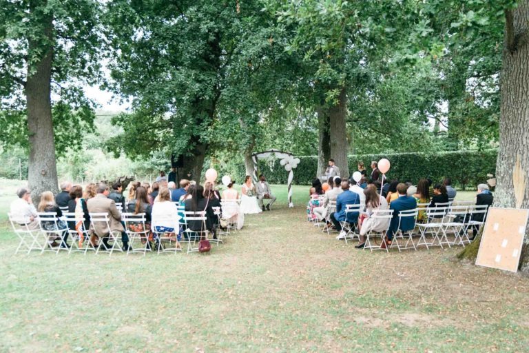 Une cérémonie de mariage émouvante et touchante se déroule en plein air, dans une zone herbeuse entourée d'arbres. Les invités sont assis sur des chaises pliantes blanches face à l'autel, où le couple se tient debout sous une arche ornée de tissu blanc. Les participants tiennent des ballons et un grand tableau est installé à droite des chaises.