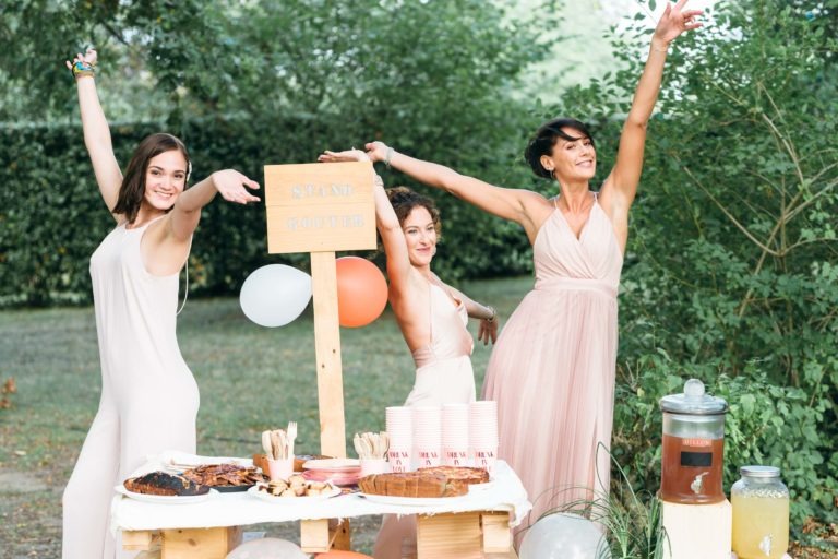 Trois femmes en robe rose pâle posent avec exubérance, les bras levés, derrière une table en bois rustique lors d'un événement en plein air. La table est ornée de produits de boulangerie, de tasses, d'un distributeur de limonade, de ballons et d'un panneau « SWEET CORNER ». L'arrière-plan est une végétation luxuriante créant une atmosphère festive et joyeuse rappelant un mariage émouvant et touchant.