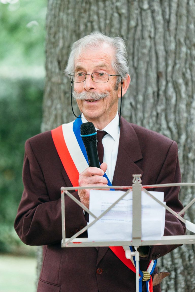 Un homme d'un certain âge, portant une moustache distinguée, des lunettes et un costume sombre orné d'une ceinture tricolore, parle dans un micro. Il se tient dehors, devant un arbre, tenant des papiers sur un pupitre devant lui, lors de ce qui semble être un mariage émouvant et touchant.