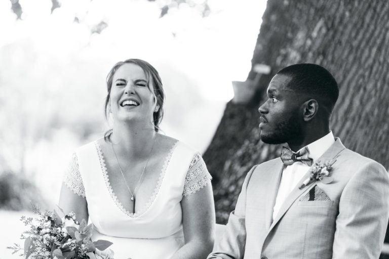 Une photo en noir et blanc montrant un couple de mariés assis à l'extérieur devant un arbre. La mariée rit joyeusement, tenant un bouquet de fleurs. Elle porte une robe blanche aux manches en dentelle. Le marié, en costume clair avec nœud papillon et boutonnière, la regarde et sourit. Un mariage émouvant et touchant magnifiquement capturé.