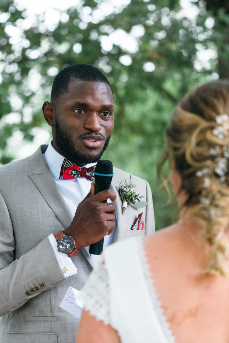 Un homme en costume gris clair avec un nœud papillon rouge et noir parle dans un micro lors d'un mariage en plein air, émouvant et touchant. Il a une boutonnière fleurie épinglée à son revers et porte une montre-bracelet. L'arrière-plan est une verdure floue. Une femme aux cheveux blonds ornés d'épingles décoratives écoute en tournant le dos à la caméra.