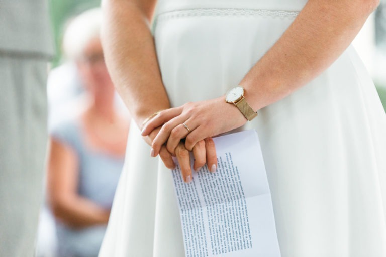 Une personne vêtue d'une robe blanche tient dans ses deux mains un morceau de papier avec un texte, peut-être un discours. Elle porte également une montre au poignet gauche et une bague à la main gauche. L'arrière-plan flou suggère un cadre intime pour ce qui pourrait être un moment de mariage émouvant et touchant.