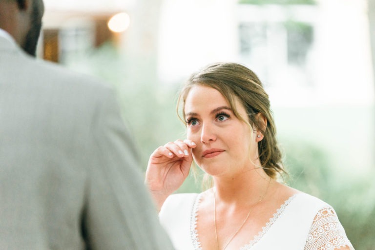 Une mariée en robe blanche aux manches en dentelle essuie une larme de son œil pendant le mariage émouvant et touchant. Ses cheveux sont coiffés en chignon lâche et elle porte un collier à pendentif en or. Le marié, vêtu d'un costume gris, se tient au premier plan, dos à la caméra. L'arrière-plan est flou.