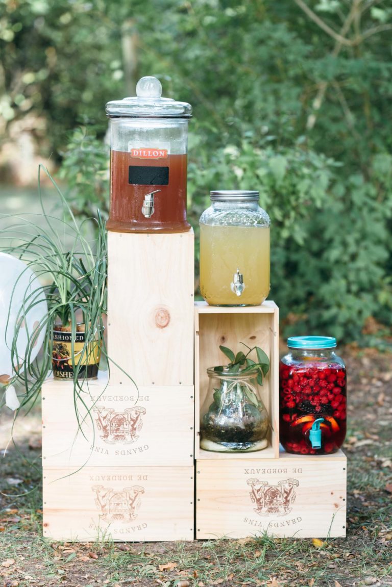 Un stand de boissons rustique en plein air comprend des caisses en bois empilées pour soutenir des distributeurs en verre. Un distributeur contient du thé glacé et un autre de la limonade. Dans un mariage émouvant et touchant, la configuration comprend également une plante dans une canette, un bocal en verre avec une plante à l'intérieur et un bocal rempli de baies rouges trempées dans du liquide.