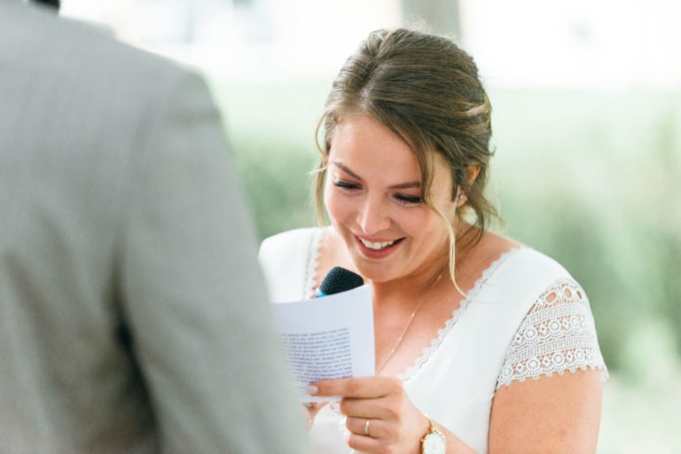 Une mariée aux cheveux châtain clair attachés en chignon lâche sourit et lit ses vœux sur un papier lors de son mariage émouvant et touchant. Elle tient un micro et porte une robe blanche à manches courtes en dentelle. Une personne en costume gris se tient à sa gauche, légèrement floue.