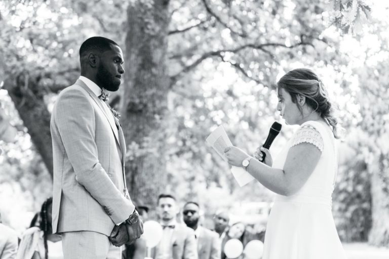 Une photo en noir et blanc capture une cérémonie de mariage émouvante et touchante en plein air, sous une voûte d'arbres. Un marié en costume clair regarde la mariée, qui lit ses vœux sur un papier tout en tenant un micro. Plusieurs invités sont assis en arrière-plan, observant attentivement le couple.