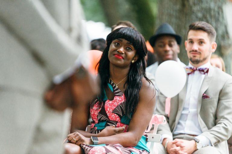 Une femme vêtue d'une robe colorée, aux cheveux longs et foncés, portant une montre connectée, sourit et écoute attentivement lors d'un événement en plein air. Elle est assise parmi d'autres personnes, dont certaines sont habillées de façon formelle. En arrière-plan de ce mariage émouvant et touchant, un homme en costume clair avec un nœud papillon tient un ballon près des arbres.