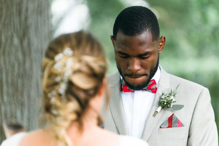 Un marié, vêtu d'un costume beige, d'un nœud papillon rouge et blanc et d'une boutonnière avec de la verdure, se tient debout face à une mariée qui a le dos tourné. Les cheveux blonds de la mariée sont coiffés en une tresse basse ornée de petites fleurs. Ils semblent être en plein air avec des arbres flous en arrière-plan lors de ce mariage émouvant et touchant.