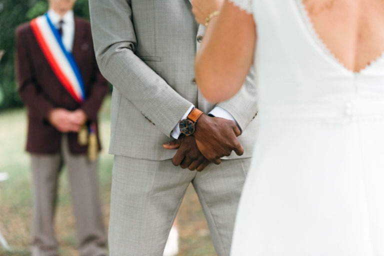 Un marié en costume gris clair et montre-bracelet marron joint ses mains lors d'un mariage émouvant et touchant. Une mariée en robe blanche avec des détails en dentelle sur les manches et le dos se tient en face de lui. Une personne en costume sombre avec une ceinture tricolore regarde en arrière-plan.