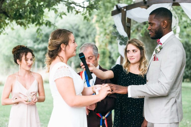 Une mariée en robe blanche et un marié en costume gris clair échangent leurs alliances sous une arche en bois décorée de tissu blanc et de verdure en plein air. Dans ce mariage émouvant et touchant, une femme aux cheveux bouclés dans une robe rose clair se tient à gauche, et une autre femme en noir tient un micro à côté d'un homme âgé.