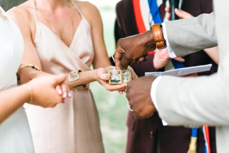 Gros plan d'un mariage émouvant et touchant où une personne en costume clair avec une montre marron tient un coffret à bijoux affichant des bagues détaillées. Une autre personne en robe claire, partiellement visible, soutient le coffret. L'officiant en arrière-plan tient des papiers et porte une ceinture à rayures colorées.