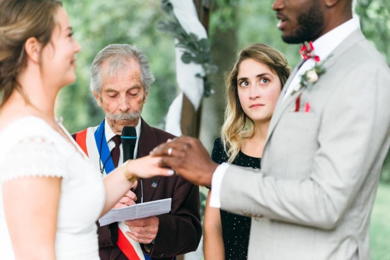 Un couple de mariés échangeant leurs alliances lors d'une cérémonie de mariage en plein air. La mariée est à gauche, partiellement visible, tenant la main du marié. L'officiant, un homme âgé aux cheveux gris et à la moustache, lit un journal. Une femme blonde en noir observe attentivement en arrière-plan ce mariage émouvant et touchant.