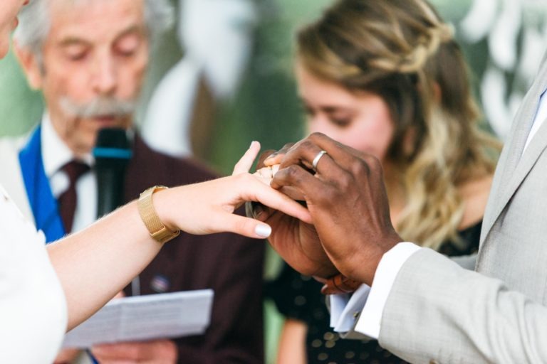 Un gros plan montre un couple échangeant des alliances lors de leur cérémonie de mariage. Le marié place une bague au doigt de la mariée, capturant l'essence de leur mariage émouvant et touchant. L'officiant, tenant un micro et quelques notes, et une demoiselle d'honneur, concentrée sur le couple, sont flous en arrière-plan.