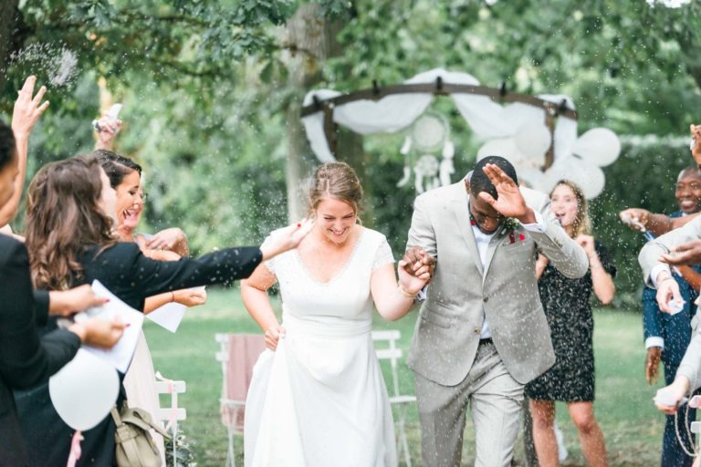 Une cérémonie de mariage joyeuse en plein air. Les mariés souriants se tiennent la main et marchent dans une allée extérieure pendant que les invités les couvrent de riz. Le mariage émouvant et touchant voit la mariée en robe blanche et le marié en costume gris clair, sur fond de verdure luxuriante et d'une arche décorée.