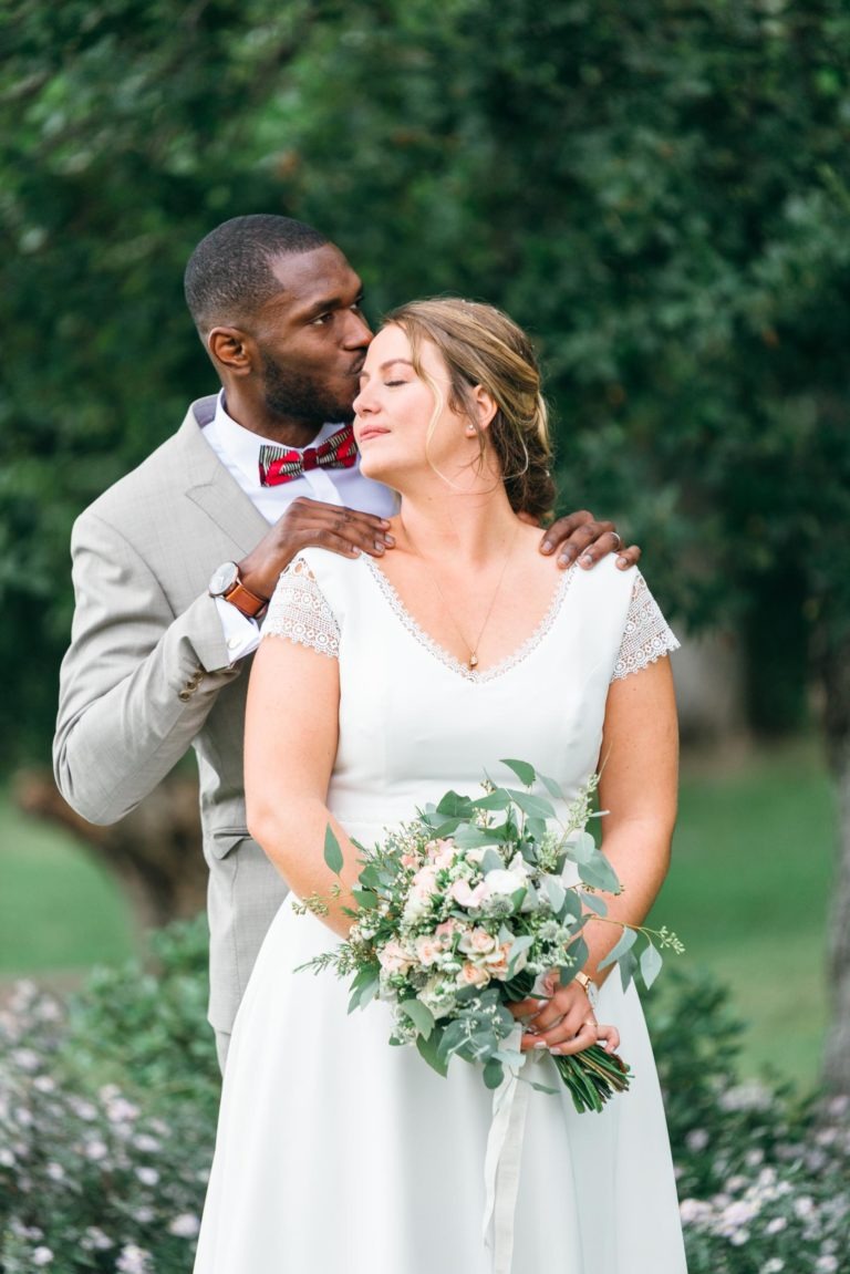 Un marié en costume gris clair embrasse une mariée en robe blanche et l'embrasse sur le front. La mariée tient un bouquet luxuriant de fleurs blanches et roses avec de la verdure. Ils se tiennent à l'extérieur sur fond d'arbres verts, partageant un mariage émouvant et touchant le jour de leur mariage.