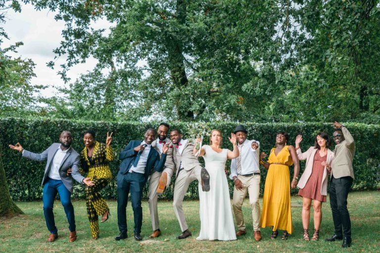 Un groupe joyeux de neuf personnes vêtues de tenues formelles et semi-formelles, dont une mariée en robe blanche et un marié en costume beige, posent ensemble à l'extérieur sous un arbre. Souriants et frappants dans des poses ludiques, ils capturent l'essence d'un événement festif, probablement un mariage émouvant et touchant.