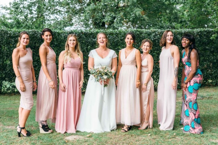 Une mariée se tient dehors devant une haie avec sept demoiselles d'honneur, créant un mariage émouvant et touchant. La mariée porte une robe blanche et tient un bouquet de fleurs. Les demoiselles d'honneur portent différents styles de robes rose poudré, à l'exception d'une robe colorée à motifs. Elles sourient ou rient toutes sous la voûte des arbres.