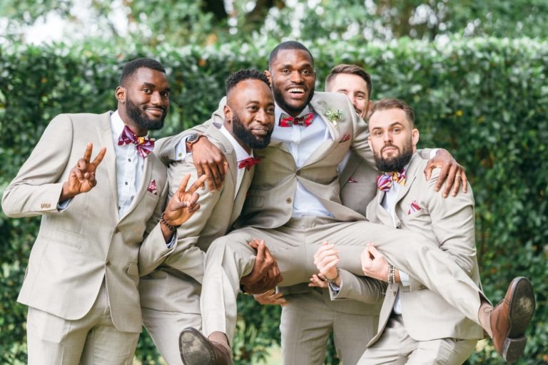 Cinq hommes en costume beige avec un nœud papillon rouge posent de manière ludique à l'extérieur. Quatre d'entre eux soutiennent le cinquième homme, qui est au centre, souriant joyeusement. Le décor est vert, suggérant un jardin ou un parc, créant une atmosphère animée et festive de mariage émouvant et touchant.