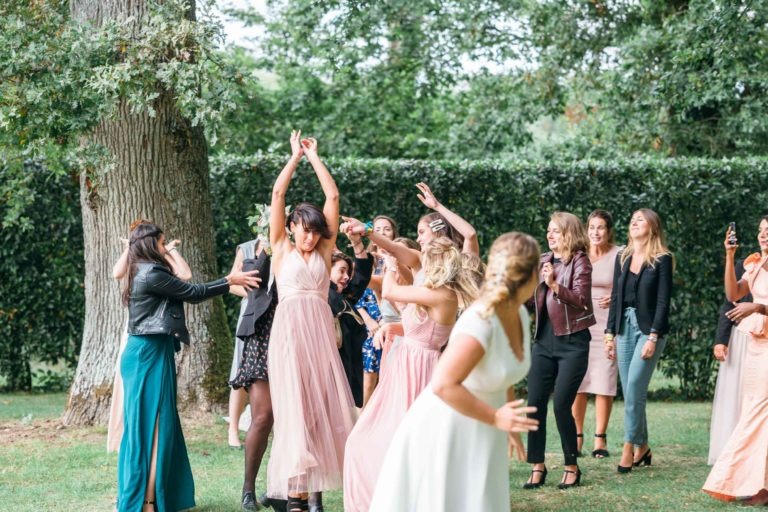 Un groupe de femmes lors d'un mariage en plein air se penchent agressivement vers le haut, essayant d'attraper un bouquet lancé par une femme en robe blanche, le dos tourné vers la caméra. Le décor comprend de grands arbres et une haie verte luxuriante. Certaines portent des robes formelles, tandis que d'autres portent des vestes par-dessus leurs tenues - un moment de mariage vraiment émouvant et touchant.