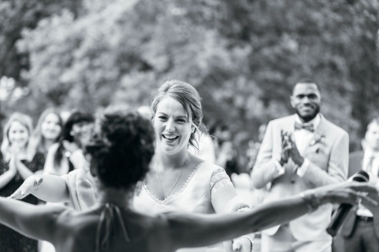 Une image en noir et blanc capture un moment de joie alors qu'une femme enlace une autre personne à bras ouverts. Autour d'elles, un groupe de personnes regarde et applaudit, ajoutant à l'ambiance de ce mariage émouvant et touchant. L'arrière-plan est flou, mettant en valeur les arbres, donnant une impression de décor extérieur. L'atmosphère semble festive et festive.