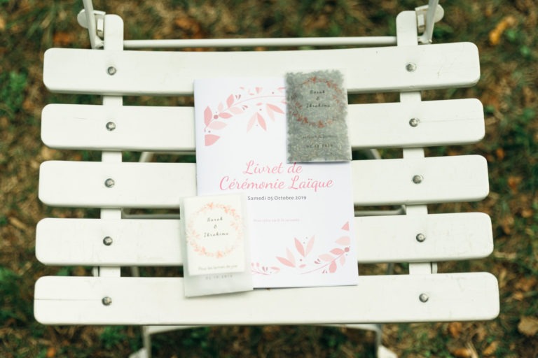 Une chaise blanche à lattes abrite trois objets : un livret rose et blanc intitulé « Livret de Cérémonie Laïque », vraisemblablement un programme de mariage, flanqué de deux petits carnets fermés aux élégants motifs floraux. L'arrière-plan révèle un sol herbeux, suggérant un mariage émouvant et touchant.