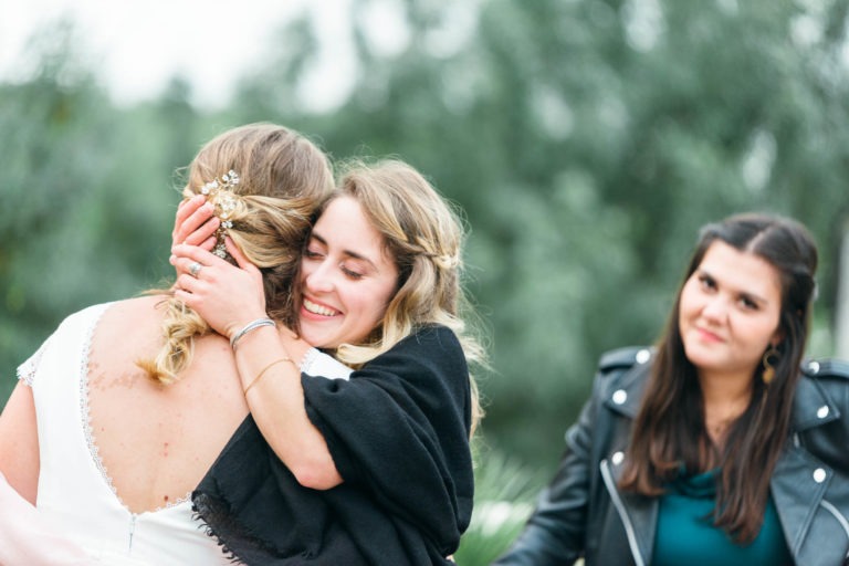 Deux femmes s'embrassent chaleureusement ; celle de gauche tourne le dos, les cheveux ornés d'une pince décorative, tandis que celle de droite sourit, les yeux fermés. Une troisième femme se tient à proximité, vêtue d'une veste en cuir noir, observant l'étreinte avec un doux sourire. L'arrière-plan, évoquant un mariage émouvant et touchant, est légèrement flouté.