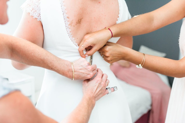 Trois personnes aident une mariée à ajuster sa robe blanche, en se concentrant sur la fixation des boutons le long du dos. La robe de la mariée est ornée de dentelles sur les manches et le dos. Dans ce mariage émouvant et touchant, les mains des assistantes, ornées de bracelets, travaillent avec attention. Un vêtement rose est visible à l'arrière-plan.