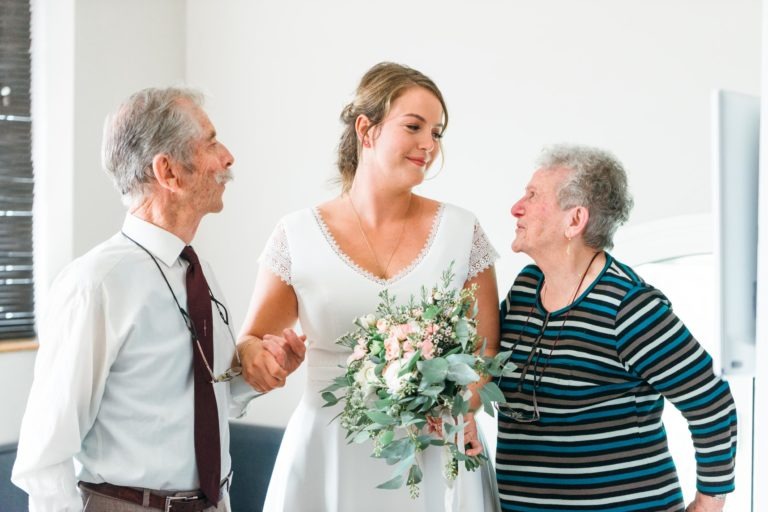 Une mariée en robe blanche tient un bouquet de fleurs entre un homme et une femme âgés. L'homme, vêtu d'une chemise blanche et d'une cravate, lui tient la main, et la femme, vêtue d'une chemise rayée, la regarde. Ils semblent tous partager un moment de joie à l'intérieur lors de ce mariage émouvant et touchant.