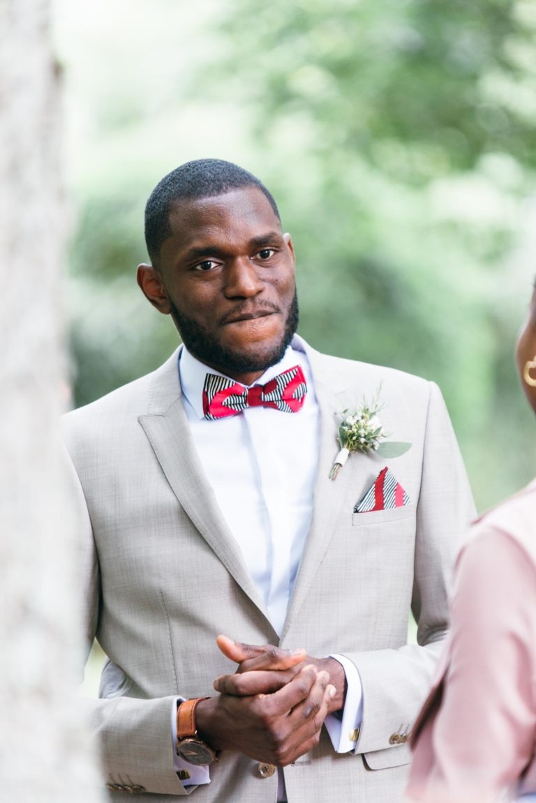 Un homme vêtu d'un costume beige clair, d'une chemise blanche et d'un nœud papillon à motifs rouges se tient debout à l'extérieur, les mains jointes. Il a une barbe soigneusement taillée et porte une boutonnière fleurie sur son revers. L'arrière-plan présente des éléments verts flous, suggérant un mariage émouvant et touchant.