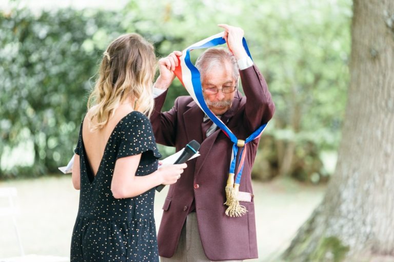 Un homme d'âge mûr, vêtu d'un blazer bordeaux et de lunettes, se tient sous un arbre tandis qu'une femme vêtue d'une robe noire à dos ouvert l'aide à enfiler une ceinture de cérémonie bleue et dorée. Le décor semble être celui d'un événement en plein air, peut-être un mariage émouvant et touchant, avec de la verdure en arrière-plan. Les deux individus sont concentrés sur la ceinture.